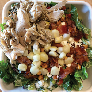 A salad in a takeout container topped with shredded chicken, bacon pieces, corn, chickpeas, feta cheese, and chopped romaine lettuce.