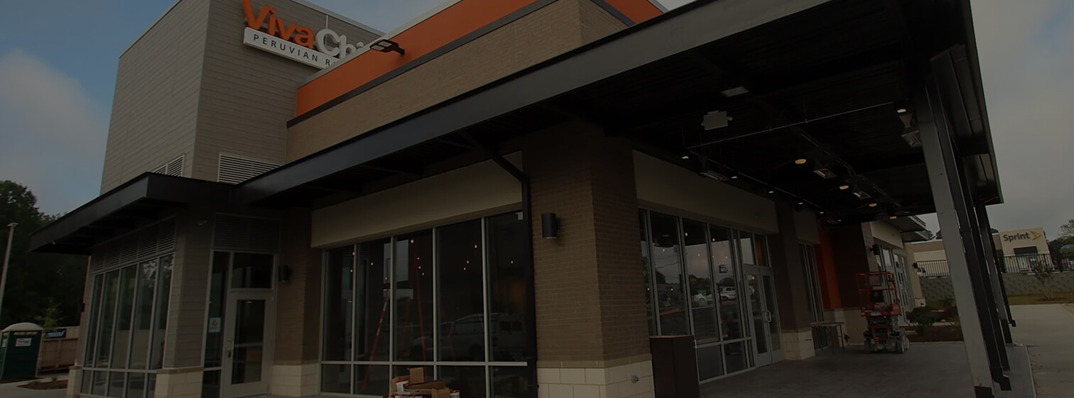 A modern commercial building with large glass windows, an orange and brown exterior, and a covered entryway. Construction equipment and materials are visible outside.