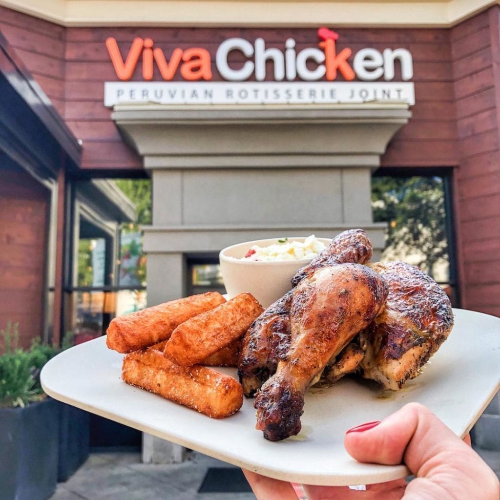 A hand holds a plate with Peruvian rotisserie chicken, fried yucca, and coleslaw in front of a Viva Chicken restaurant, with the restaurant sign clearly visible in the background.