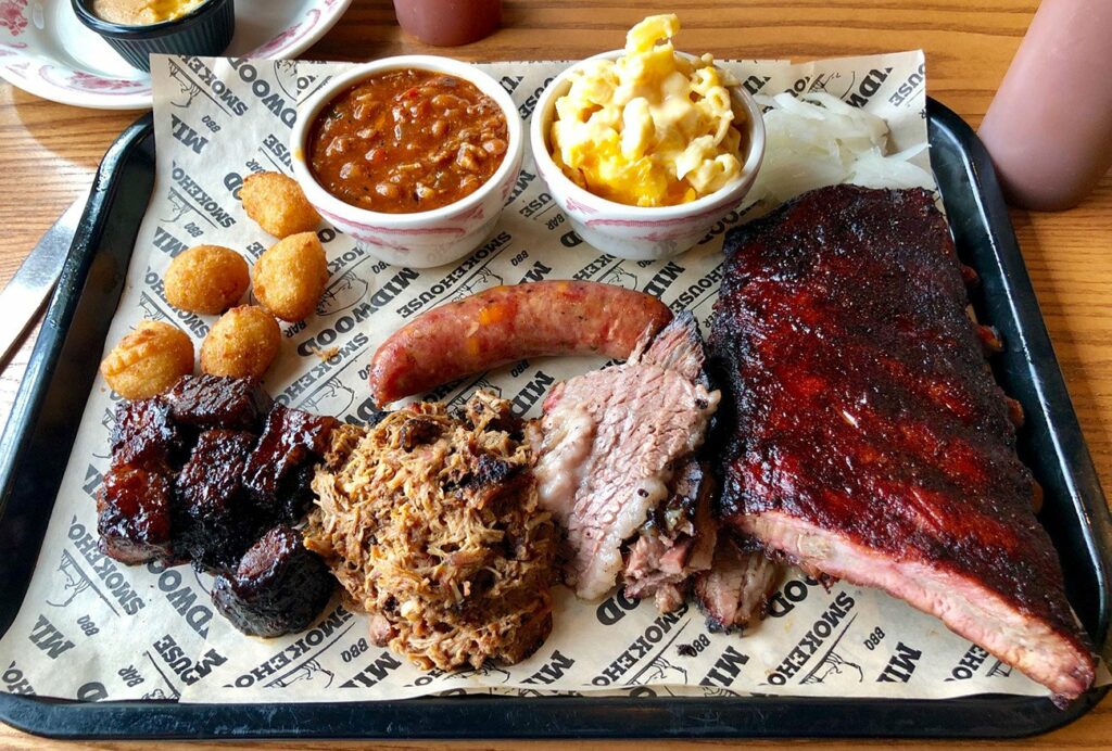 A tray with barbecue foods including ribs, brisket, pulled pork, sausage, burnt ends, hush puppies, baked beans, macaroni and cheese, and a small portion of onions on branded parchment paper.