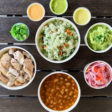 A top-down view of bowls containing grilled chicken, green rice with vegetables, pinto beans, guacamole, pickled onions, chopped green onions, and three different sauces on a wooden table.