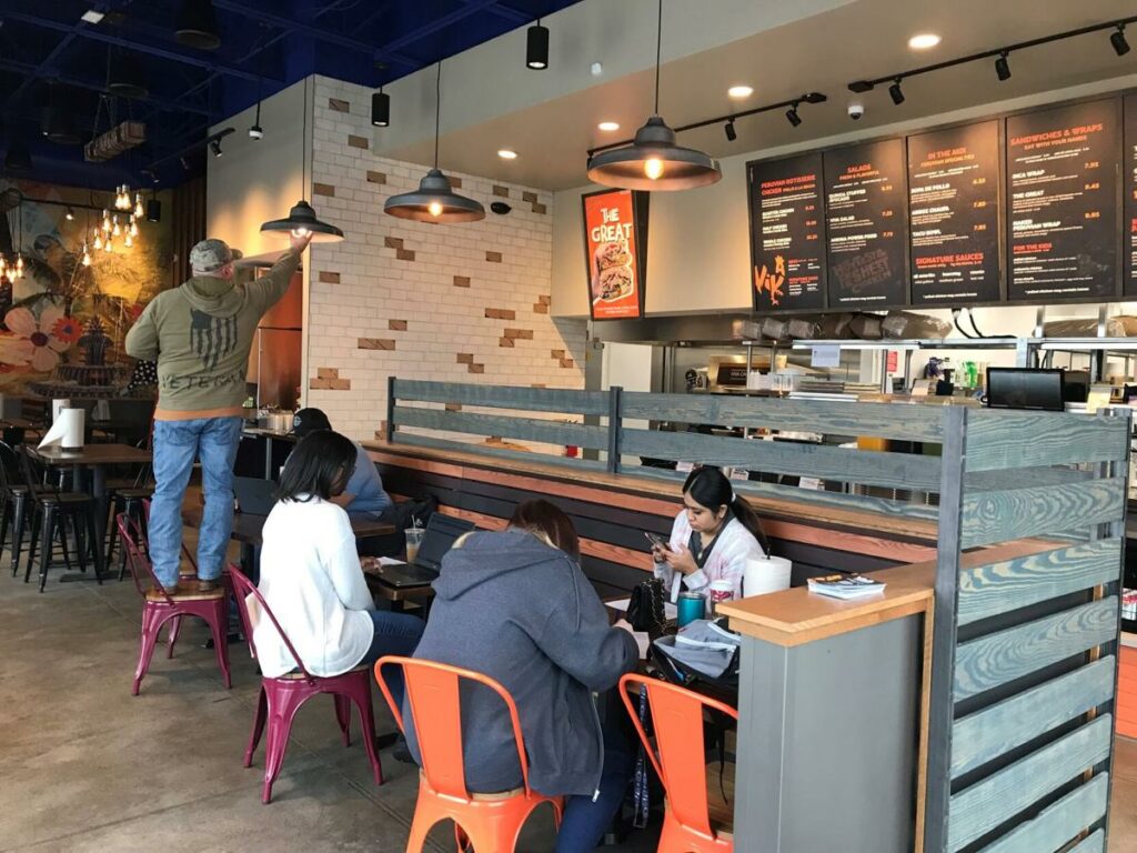 People sit at tables in a casual restaurant with a modern industrial design. One person stands on a chair adjusting something on the wall, while others eat or look at their phones. The menu is displayed above the counter.
