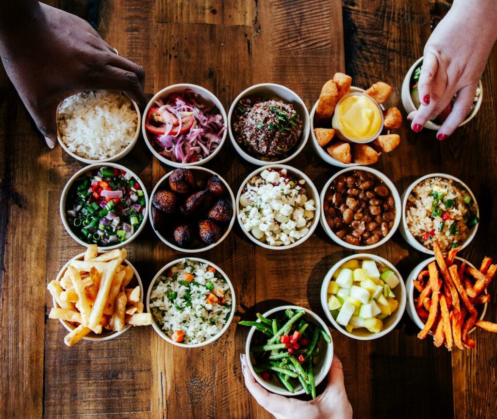 Three hands reach for an assortment of small bowls filled with various foods, such as rice, beans, fries, salads, fried plantains, sauces, and pickled vegetables, arranged on a wooden table.