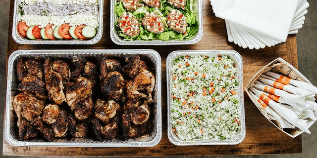 A variety of catered foods on a wooden table, including grilled chicken, rice with herbs and vegetables, salad with tomatoes and cucumbers, green salad with dressing, napkins, and wrapped cutlery.