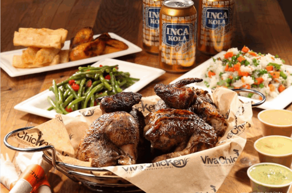 A basket of roasted chicken pieces with Viva Chicken branding, served with green beans, yucca fries, rice with vegetables, dipping sauces, and cans of Inca Kola soda on a wooden table.