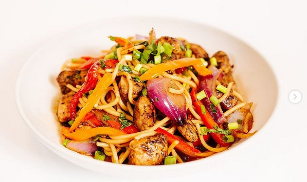 A white bowl filled with stir-fried noodles, grilled chicken, sliced red onions, bell peppers, and carrots, garnished with chopped herbs and green onions, against a plain white background.