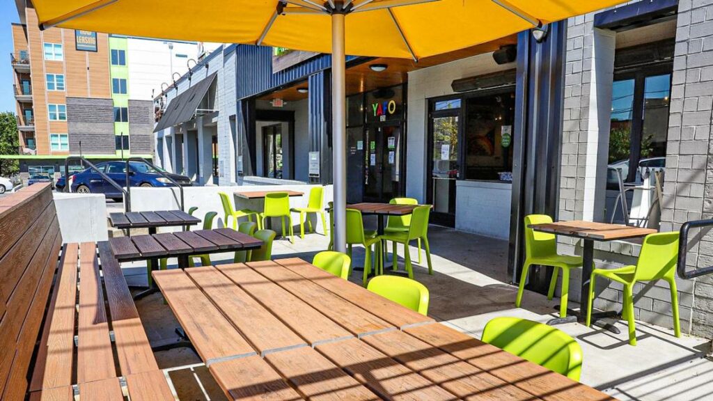 Outdoor patio seating at a restaurant with wooden tables, green chairs, and a large yellow umbrella; modern building and parked cars visible in the background on a sunny day.