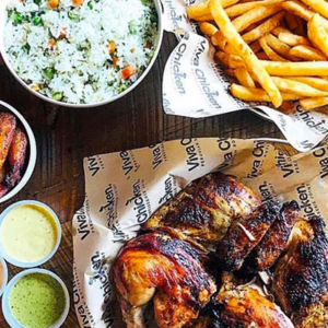 A table with roasted chicken, French fries, a bowl of rice with vegetables, chicken wings, and three dipping sauces. Food is served on branded paper labeled 