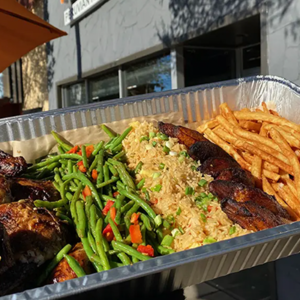 A takeout tray filled with grilled chicken, sautéed green beans with red peppers, fried rice, fried plantains, and a serving of French fries, held up outdoors in front of a restaurant.