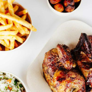 A plate of grilled chicken with sides of golden fries, fried plantains, and a bowl of rice mixed with vegetables, all served on a white table.