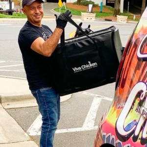 A smiling man in a black t-shirt and cap carries a large Viva Chicken delivery bag while standing next to a colorful vehicle on a street corner.