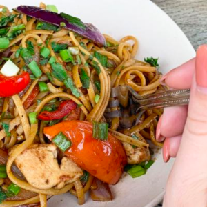 A hand holding a fork is about to pick up stir-fried noodles with chicken, red bell peppers, green onions, and herbs served on a white plate.