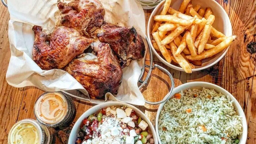 A wooden table with plates of grilled chicken, French fries, seasoned rice with vegetables, a salad topped with feta cheese, and two small containers of dipping sauces.