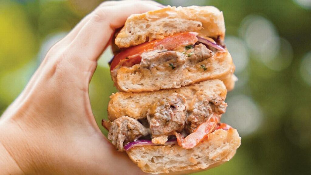 A hand holds a thick sandwich filled with chunks of meat, sliced tomato, and fresh greens between two pieces of rustic bread, with a blurred green background.
