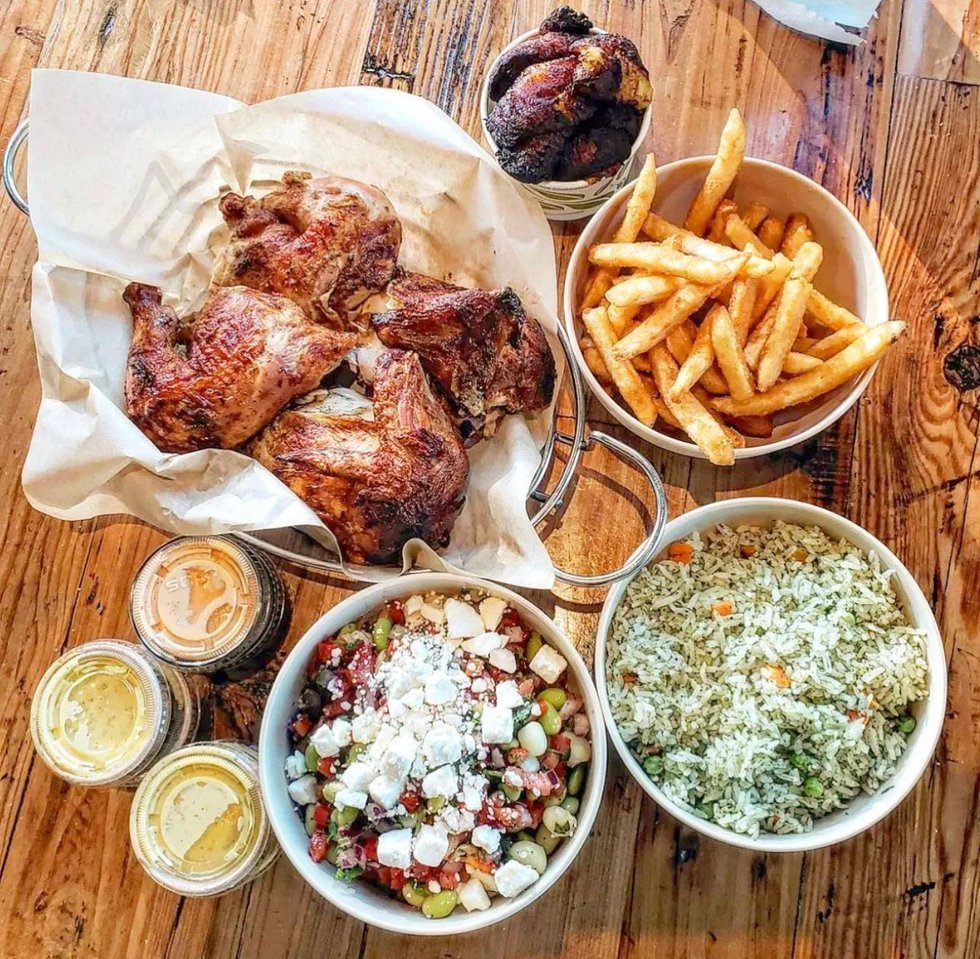 A wooden table set with roasted chicken pieces, a bowl of French fries, a bowl of yellow rice with vegetables, a bowl of Greek salad with feta cheese, small containers of sauce, and grilled chicken wings.