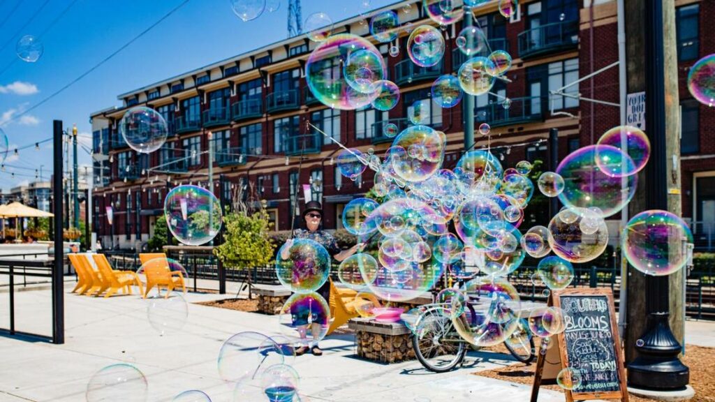 A street scene with colorful soap bubbles floating in the air, a person standing near a bike, yellow chairs, plants, and apartment buildings in the background under a blue sky.