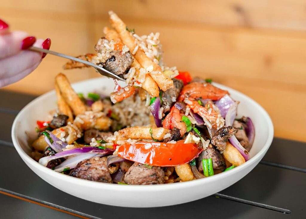 A hand holding a fork lifts a bite of food from a bowl filled with fries, grilled beef, red onions, tomatoes, rice, and herbs. The dish is colorful and served on a dark table.