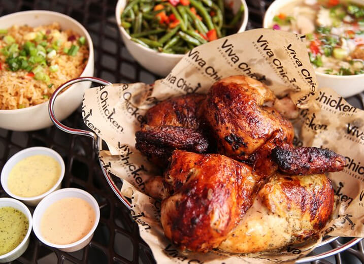 A basket of roasted chicken pieces on Viva Chicken paper is surrounded by sides of rice, green beans, soup, and three cups of dipping sauces on a dark table.