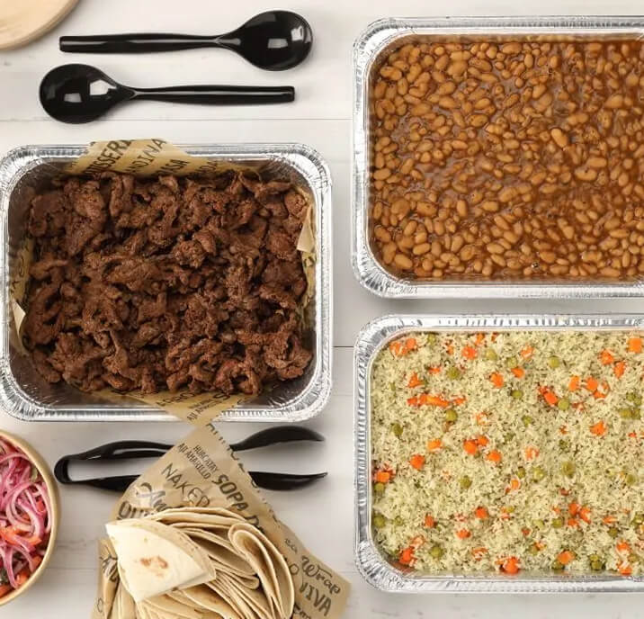 Three foil trays with grilled beef, baked beans, and rice with vegetables. Nearby are tortillas, a bowl of pickled onions, and disposable black utensils on a white table.